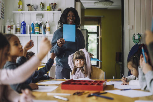 Fröhliche Lehrerin zeigt Schülern farbiges Papier, während sie sie in der Kindertagesstätte unterrichtet - MASF34617