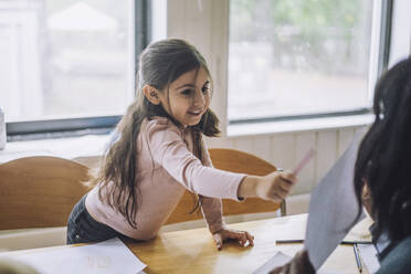 Smiling girl pointing at teacher showing drawing during art class in kindergarten - MASF34624