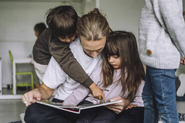 Junge, der auf ein Bilderbuch zeigt, während ein männlicher Lehrer ihm im Kindergarten eine Geschichte erzählt - MASF34629