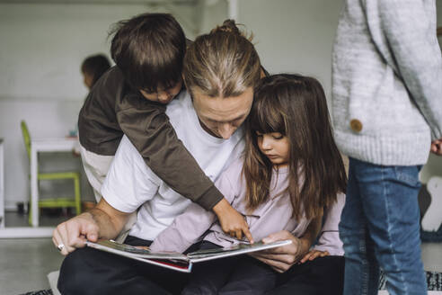 Junge, der auf ein Bilderbuch zeigt, während ein männlicher Lehrer ihm im Kindergarten eine Geschichte erzählt - MASF34629