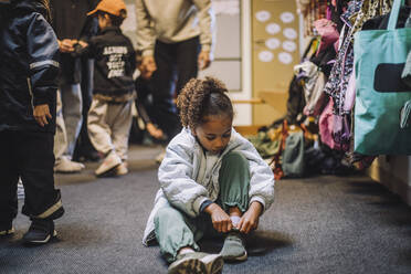 Mädchen trägt Schuh, während sie auf dem Teppich in der Kindertagesstätte sitzt - MASF34630