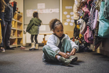 Mädchen trägt Schuh, während sie auf dem Teppich im Kindergarten sitzt - MASF34631