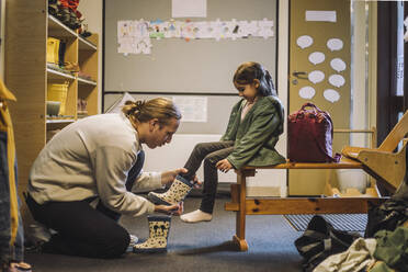 Männlicher Lehrer hilft Mädchen beim Anziehen von Gummistiefeln, während sie in der Kindertagesstätte sitzen - MASF34632
