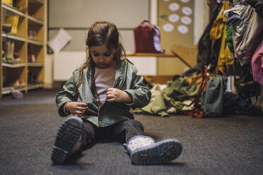 Mädchen mit Jacke sitzt auf Teppich in Kindertagesstätte - MASF34633