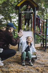 Fröhliches Mädchen gibt männlichem Lehrer High-Five, während sie auf dem Spielplatz auf der Rutsche spielt - MASF34635