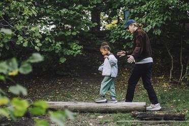 Seitenansicht eines Mädchens, das auf einem Baumstamm balanciert, während ein männlicher Lehrer sie im Park führt - MASF34636