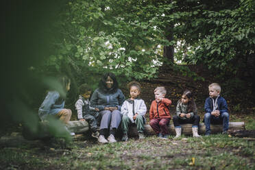 Lehrerin sitzt mit Vorschulkindern im Park auf einem Baumstamm - MASF34639