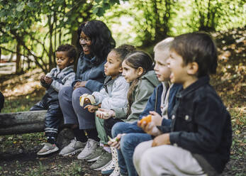 Gemischtrassige Jungen und Mädchen essen Obst, während sie mit einer Lehrerin im Park sitzen - MASF34641