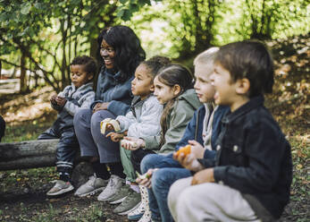 Multiracial boys and girls eating fruits while sitting with female teacher in park - MASF34641