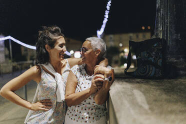 Young woman standing with arm around senior friend by wall at night - MRRF02532