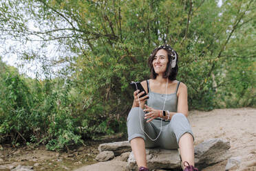 Happy young woman wearing headphones listening to music at park - MRRF02558