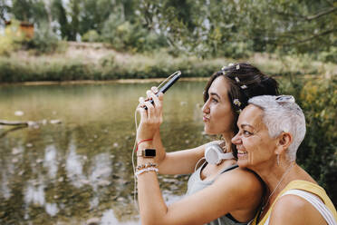 Junge Frau mit Freund spricht Selfie durch Handy am Seeufer - MRRF02560