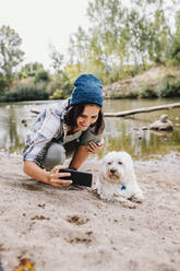 Lächelnde junge Frau macht Selfie mit Hund am Seeufer im Park - MRRF02572