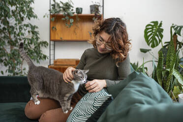 Woman stoking cat sitting on couch at home - VRAF00021