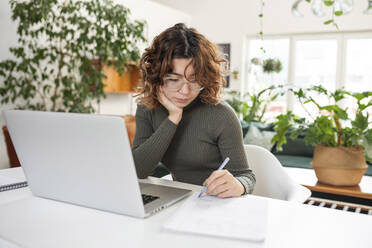 Freelancer writing notes sitting in front of laptop at home - VRAF00022