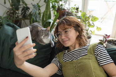 Lächelnde Frau nimmt Selfie mit Katze auf Couch zu Hause - VRAF00023