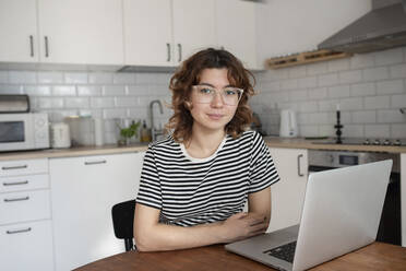 Young freelancer sitting with laptop at table in kitchen - VRAF00024