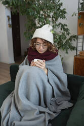 Woman wrapped in blanket sitting with cup of tea on sofa at home - VRAF00051