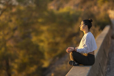 Woman sitting in lotus position practicing yoga at sunset - LJF02453