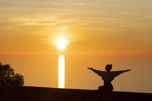 Carefree woman with arms outstretched sitting on wall at sunset - LJF02456