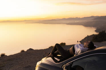 Woman with hands behind head relaxing on hood of car - LJF02462