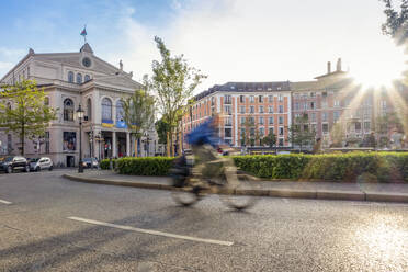 Germany, Bavaria, Munich, Blurred motion of cyclist passing through Gartnerplatz square at sunset - MAMF02485