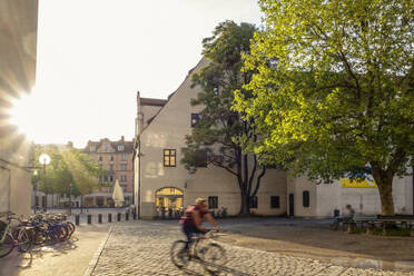 Germany, Bavaria, Munich, Cyclist riding along cobblestone street at sunset - MAMF02486