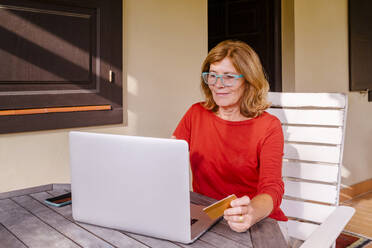 Ältere Frau beim Online-Shopping mit Kreditkarte und Laptop auf einer Veranda - MMPF00583