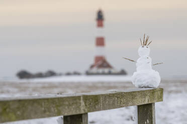 Deutschland, Schleswig-Holstein, Westerhever, Kleiner Schneemann auf einem Holzzaun mit Leuchtturm Westerheversand im Hintergrund - KEBF02561