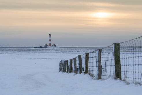 Deutschland, Schleswig-Holstein, Westerhever, Schneebedecktes Feld bei Sonnenuntergang mit Leuchtturm Westerheversand im Hintergrund - KEBF02563