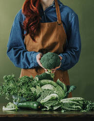 Woman wearing apron holding raw broccoli at table - VSNF00359