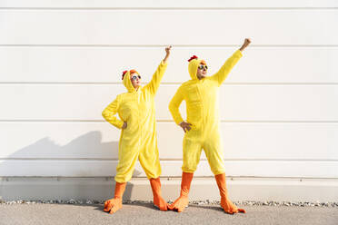 Friends wearing chicken costumes standing with arms raised in front of wall - OIPF02870