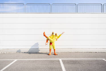 Man wearing chicken costume carrying woman in front of white wall - OIPF02882