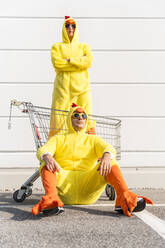 Frau im Hühnerkostüm steht in einem Einkaufswagen mit einem Mann, der auf dem Gehweg sitzt - OIPF02898