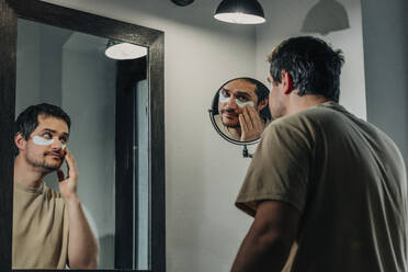 Man applying under eye patches in front of mirror - VSNF00367