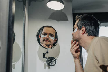 Tired man with under eye patches looking in mirror at bathroom - VSNF00368