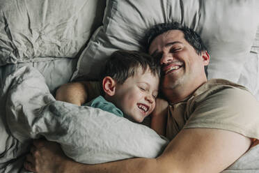 Happy father and son lying on bed in bedroom - VSNF00378