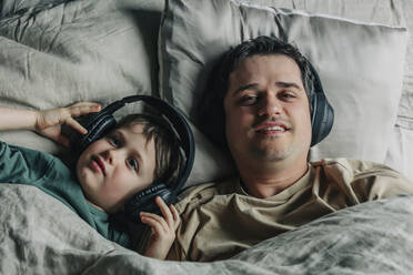 Father and son wearing headphones listening to music on bed - VSNF00379