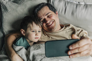 Father and son taking selfie using smart phone on bed - VSNF00380