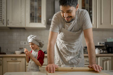 Smiling father with rolling pin by son in kitchen at home - ANAF00881