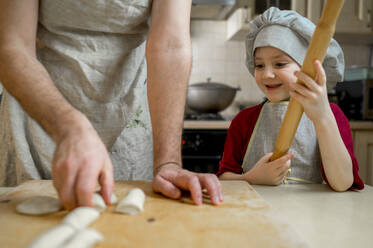 Lächelnder Junge mit Nudelholz, der seinen Vater bei der Zubereitung von Teig auf dem Küchentisch beobachtet - ANAF00883
