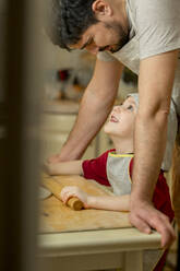 Lächelnder Junge mit Nudelholz, der seinen Vater anschaut und Essen in der Küche zubereitet - ANAF00884