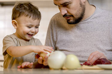 Happy boy taking white onion from father in kitchen at home - ANAF00891