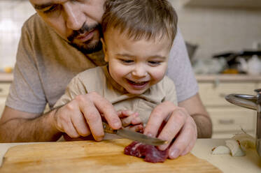 Father cutting red meat with son in kitchen - ANAF00894