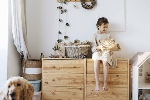 Smiling girl holding gift sitting on cabinet at home - SSYF00060