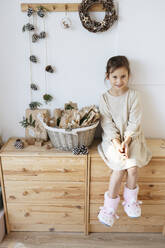 Lächelndes süßes Mädchen sitzt auf Schrank von Adventskalender Korb zu Hause - SSYF00062