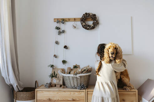 Girl hugging cocker spaniel dog sitting on cabinet at home - SSYF00063