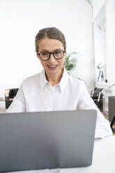 Happy businesswoman wearing eyeglasses using laptop in office - VPIF07798