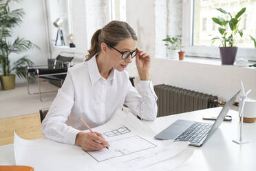 Businesswoman with blueprint looking in laptop at desk in office - VPIF07807