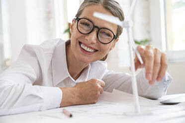 Happy mature businesswoman touching wind turbine on table in office - VPIF07814